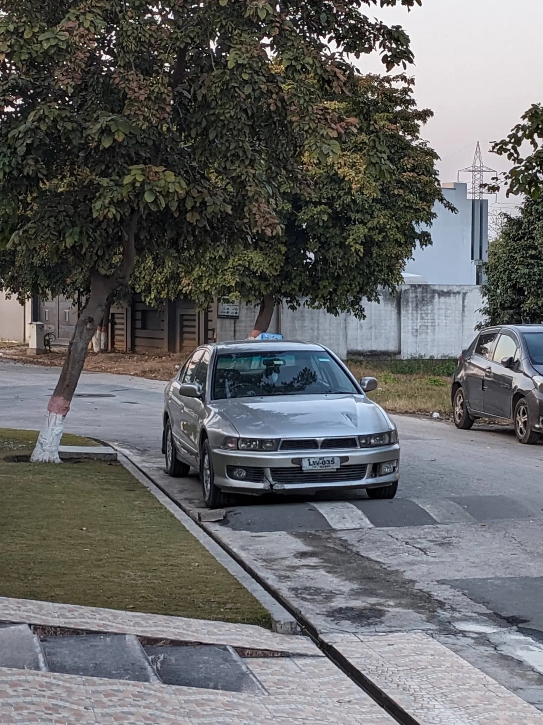 A silver Mitsubishi Galant parked on a quiet residential street, under a tree whose trunk is painted white.