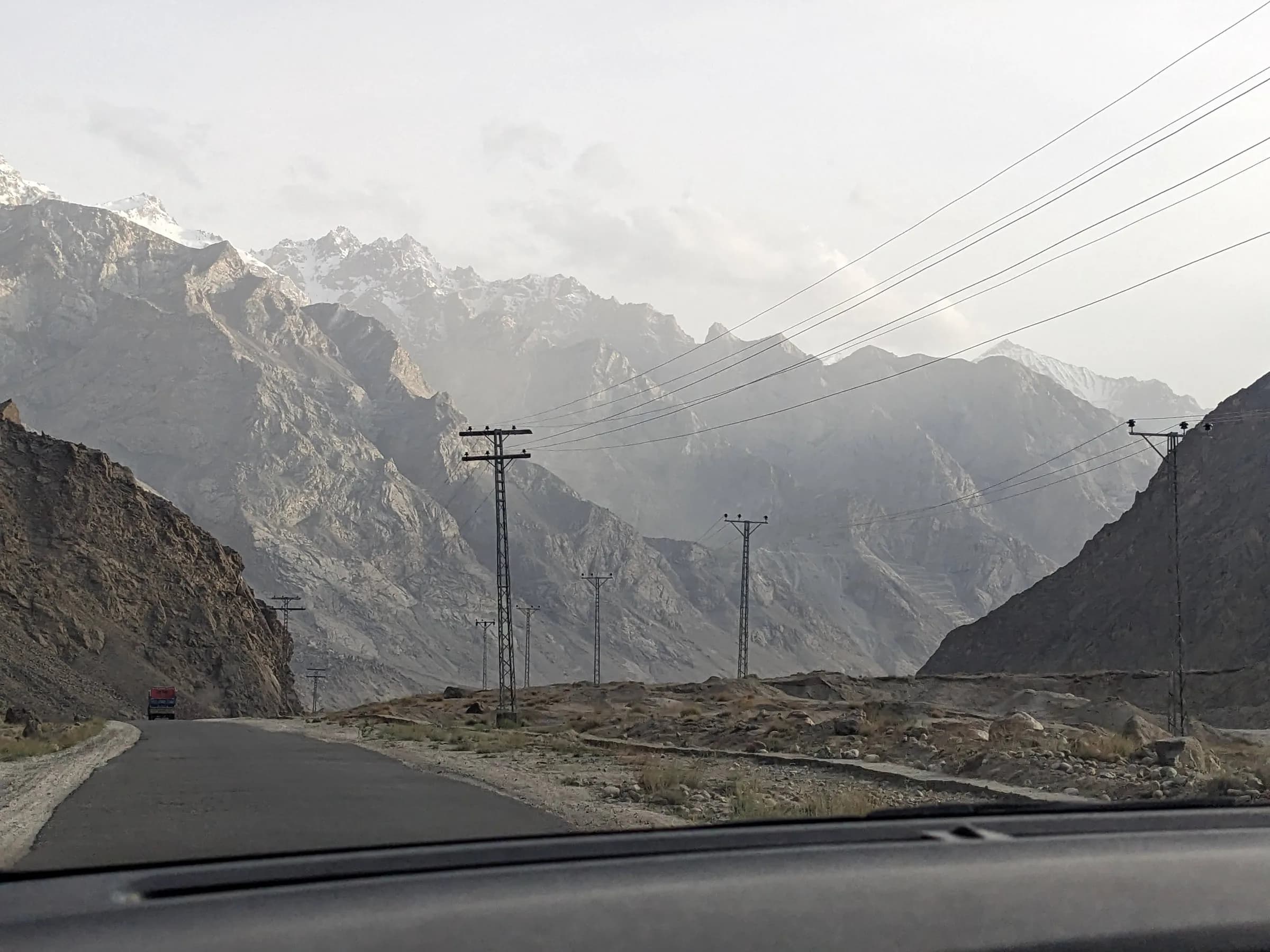 The road back to Skardu from Shigar, winding between bare rocky mountains, shot through a car windscreen with snow on the far peaks and power lines following the road.
